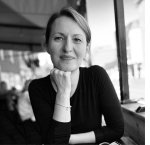 A black-and-white photo of Maja Djikic of the University of Toronto. She sits at a table with one arm on the table and the other supporting her chin. She is wearing a delicate braclet on the wrist under her chin, as well as a v-neck long-sleeved black sweater. Her slight smile seems friendly and amused, and her straight side-parted hair is pulled back from her face. There is bank of windows to one side lighting up the space behind her. 