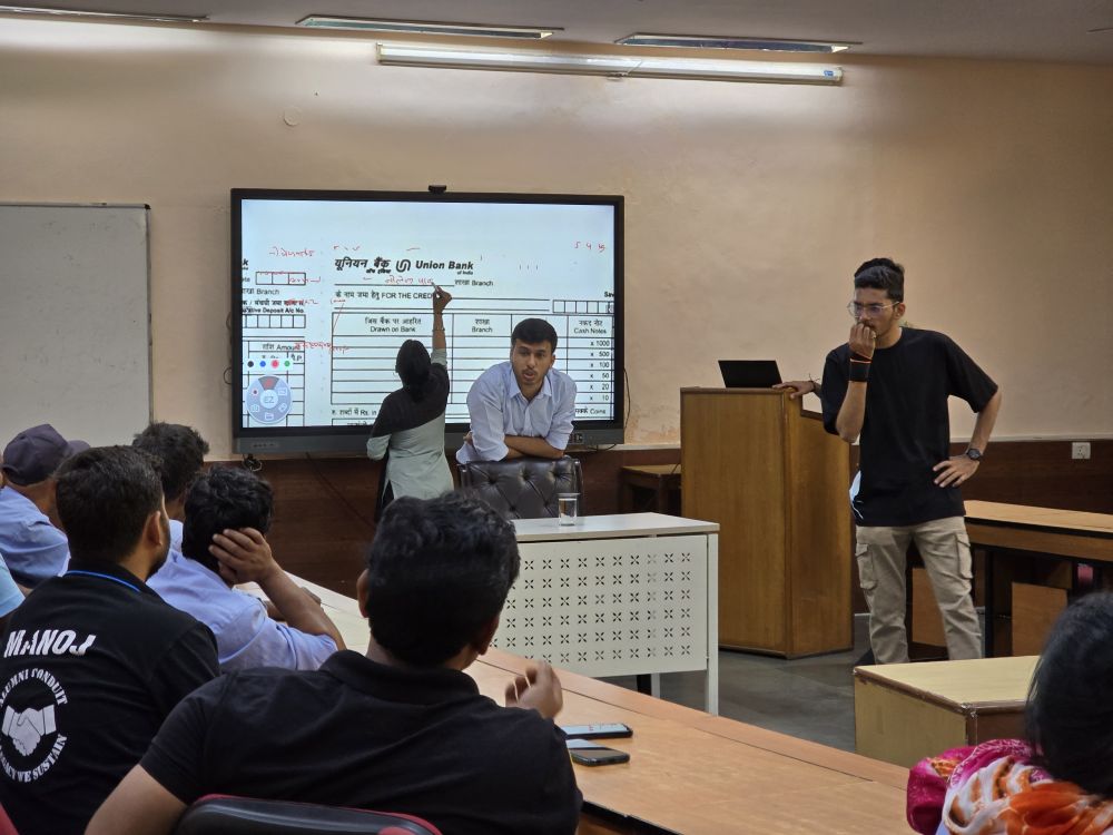 Two BIMTECH MBA students stand in a classroom, center and right of the image, as one in a light blue collared shirt leans on a black leather chair pulled under a white desk and the other stands to the right in a dark t-shirt and khaki pants looking thoughtful as he has one hand on his mouth and chin and his other on his hip. Both are speaking to about six BIMTECH staff members who are taking part in the school's Saksharta Abhiyan (literacy mission) course and seated at a table at the left. Another classmate stands behind the instructors as she practices filling out bank form displayed very large on a classroom whiteboard.